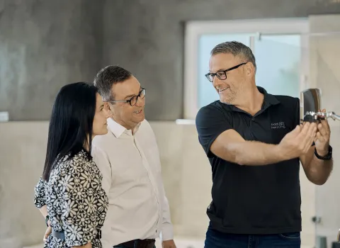 Ein Fachmann präsentiert einem interessierten Paar eine Dusche in einer Badausstellung. Alle drei lächeln, während sie die hochwertigen Produkte begutachten.