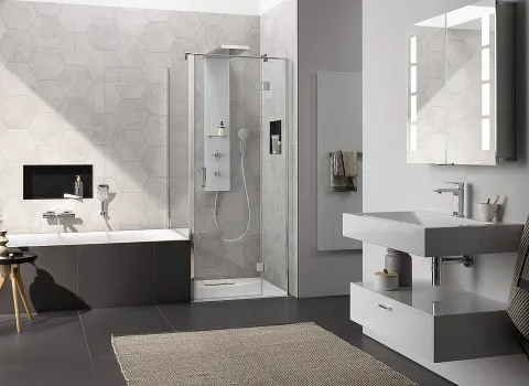 Großes Badezimmer mit einer HSK Aperto Duschkabine aus Glas, die auf grauen Sechseckfliesen montiert ist. Neben der Dusche steht eine Einbaubadewanne mit dunkler Verkleidung.