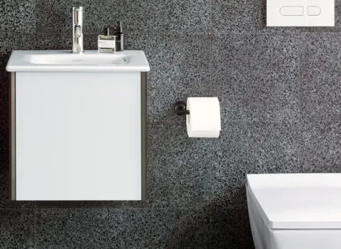 Kleines Badezimmer mit weißem Duravit Hängewaschbecken und rechteckiger Toilette vor einer dunklen strukturierten Wand. 