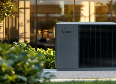 Eine Solvis-Wärmepumpe steht im begrünten Außenbereich vor einem modernen Gebäude, umgeben von Pflanzen und warmem Sonnenlicht.