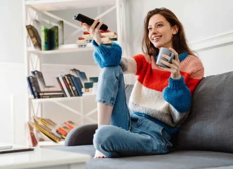 Junge Frau sitzt entspannt auf dem Sofa, hält eine Fernbedienung und genießt ihren Kaffee im Wohnzimmer.