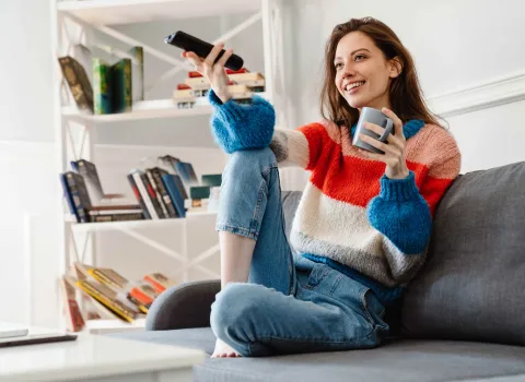 Junge Frau sitzt entspannt auf dem Sofa, hält eine Fernbedienung und genießt ihren Kaffee im Wohnzimmer.
