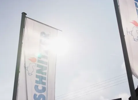 Ausschnitt eines Videos von Schnitzer TV, das Fahnen mit dem Schnitzer-Logo vor blendendem Sonnenschein zeigt.