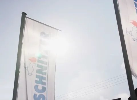 Ausschnitt eines Videos von Schnitzer TV, das Fahnen mit dem Schnitzer-Logo vor blendendem Sonnenschein zeigt.