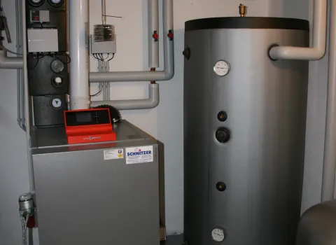 Ein moderner Heizungsraum mit grauem Warmwasserspeicher und angeschlossener Heizungsanlage ist kompakt in einer Kellerecke installiert.