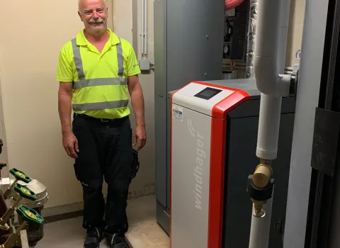 Ein Installateur in neongelbem Arbeitsshirt mit Reflexstreifen steht in einem Heizungsraum neben einer modernen Windhager-Heizungsanlage mit roter Frontverkleidung.