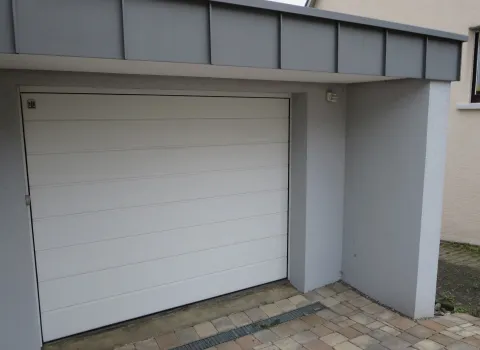 Eine moderne Einzelgarage mit weißem Sektionaltor ist in die helle Fassade eines Wohnhauses integriert.