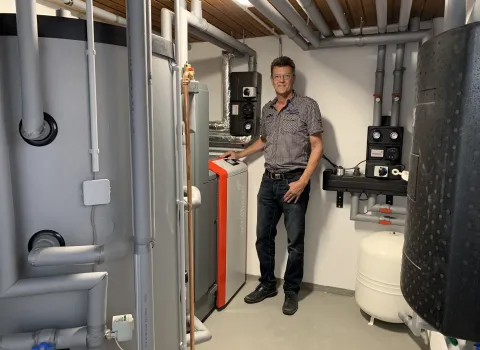 Ein Mann steht in einem modernen Heizungsraum neben einer Windhager-Heizungsanlage mit roter Front und integriertem Display.