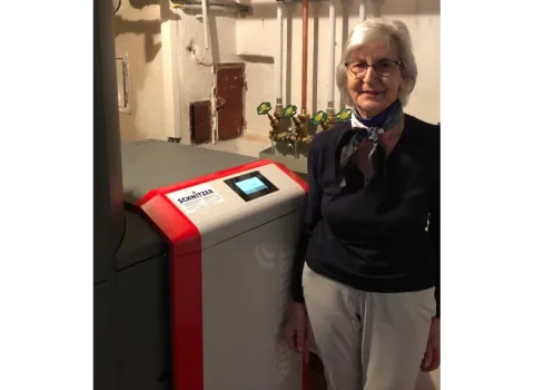 Eine ältere Frau steht in einem Heizungsraum neben einer modernen Windhager-Heizanlage mit roter und grauer Front sowie integriertem Display.