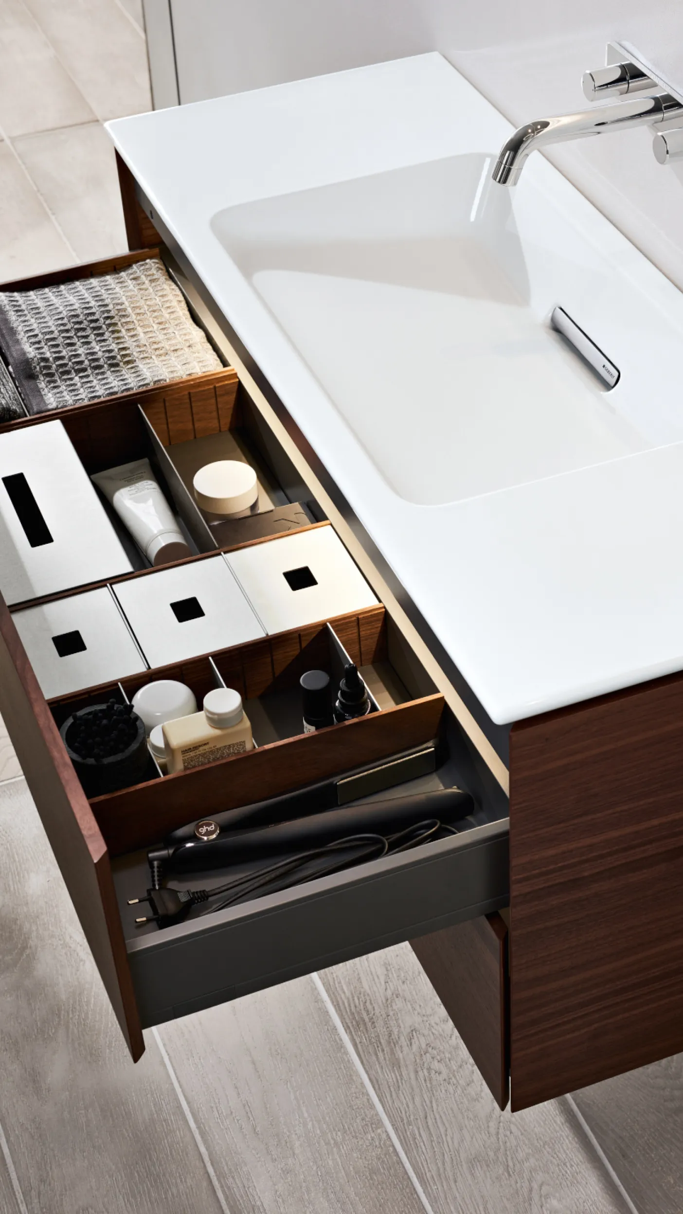 Eine geöffnete Schublade unter einem Waschbecken zeigt eine sorgfältige Anordnung von Kosmetikprodukten und Pflegeartikeln in Fächern.