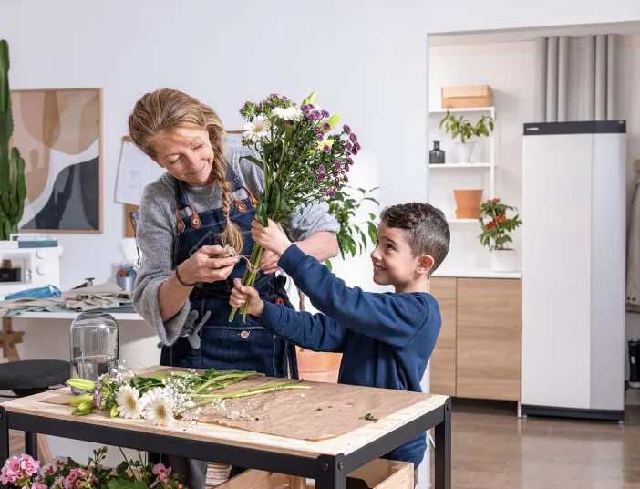 Ein kleiner Junge hilft einer Frau beim Zusammenstellen eines frischen Blumenstraußes, während sie mit einer Schere die Stiele kürzt. Die Wärmepumpen-Inneneinheit von NIBE im Hintergrund passt zur gemütlichen Atmosphäre des hellen Raumes.