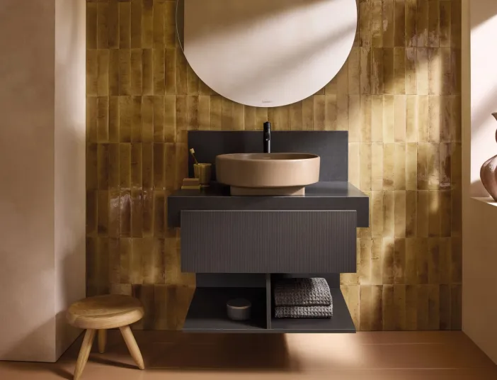 Forntansicht eines Waschtischs der Serie Balcoon von Duravit mit ockerfarbenen Aufsatzbecken und schwarzen Unterschrank vor einer goldbraun gefliesten Wand.