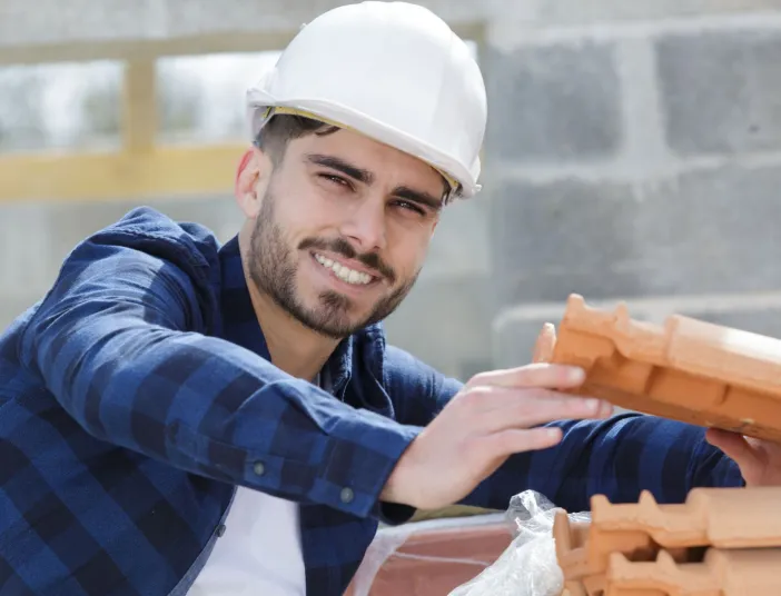 Ein junger Dachdecker mit weißem Helm posiert auf einer Baustelle mit einer Ziegel.
